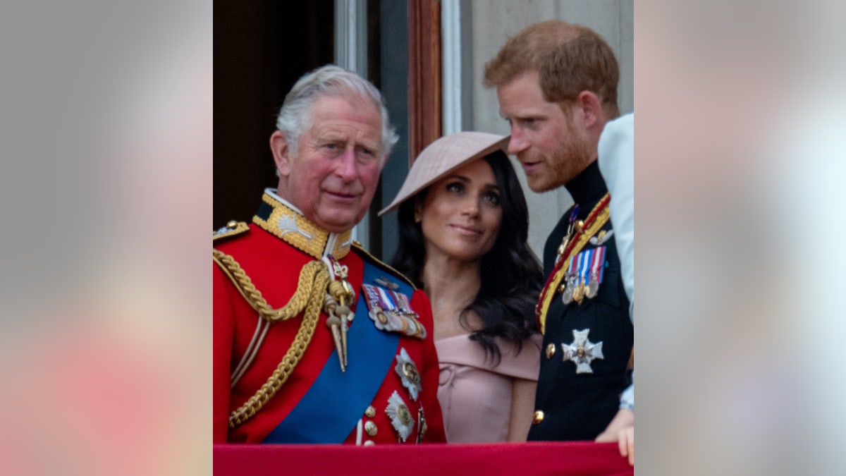 King Charles in a red suit looking away as Meghan Markle in a pink dress looks up at Prince Harry who is wearing a uniform with medals