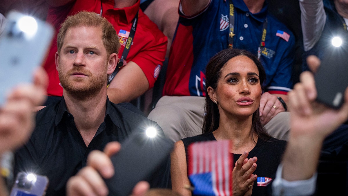 Meghan Markle and Prince Harry sitting in a crowd surrounded by cell phones