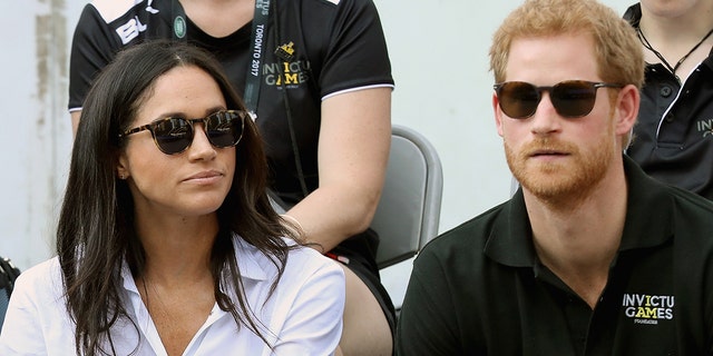Meghan Markle wearing a white blouse with black sunglasses sitting next to Prince Harry in a black shirt and sunglasses