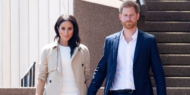 Meghan Markle wearing a white dress with a beige trench coat next to prince harry in a navy blazer and white shirt
