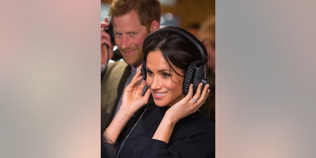 Meghan Markle in a black dress smiling and holding her headphones