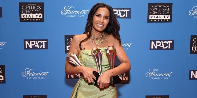 Padma Lakshmi wearing a light dress sleeveless dress holding a bunch of awards