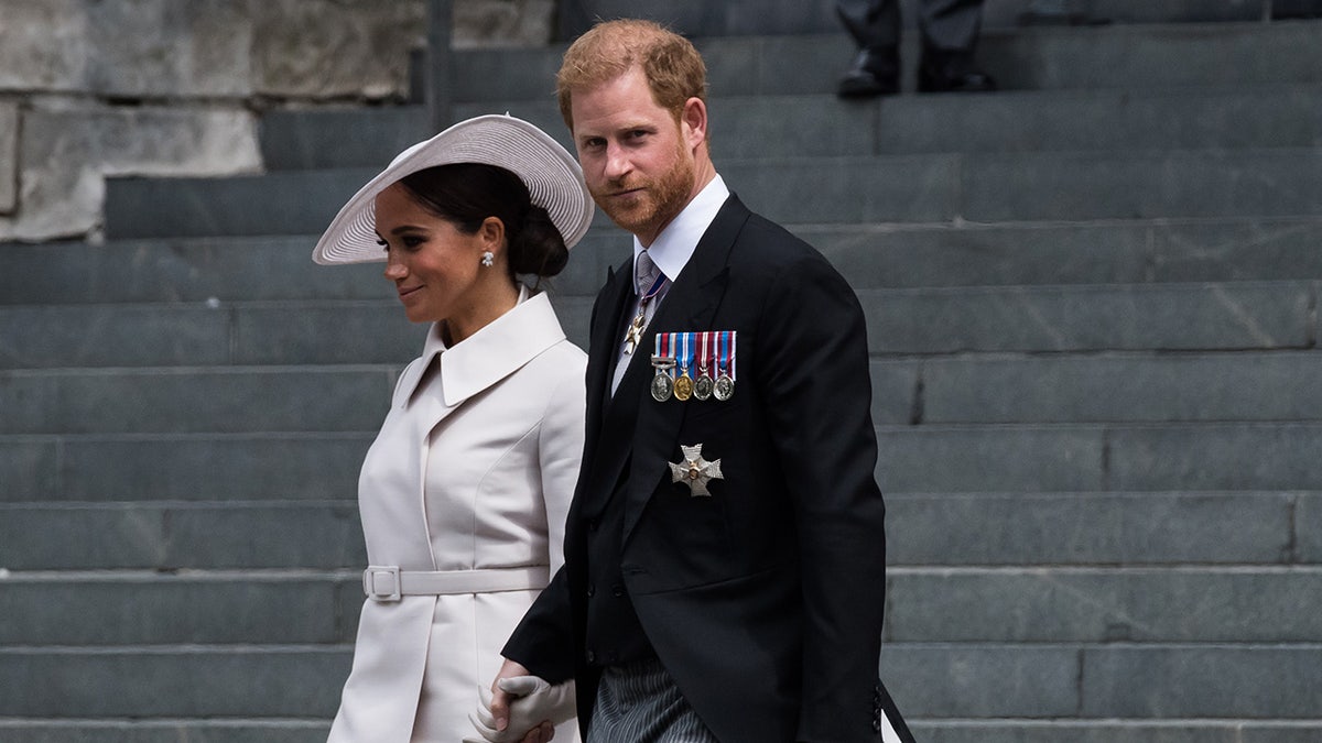 Prince Harry in a black suit with medals holding Meghan Markles hand, who is wearing a white coat dress and a matching hat