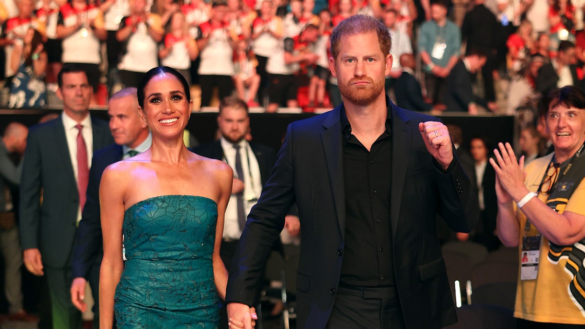 Prince Harry has his arm up in a navy suit next to Meghan Markle in a green tube dress