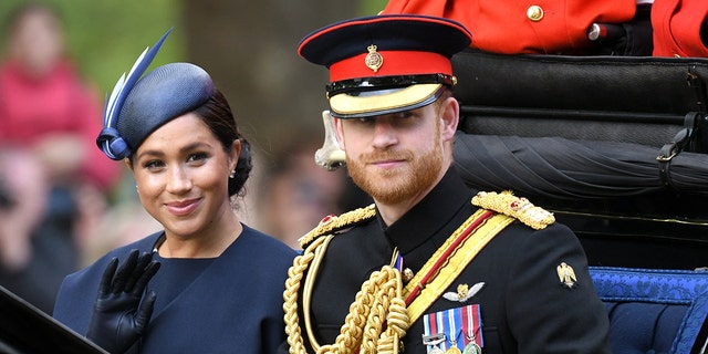 Meghan Markle in a blue dress and matching hat sitting next to Prince Harry in his military uniform