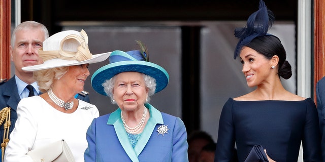 Camilla wearing a white dress chatting with Meghan Markle in a dark blue dress as Queen Elizabeth, wearing a multilayered blue dress, looks straighforward