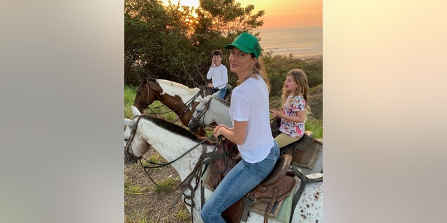 Gisele Bündchen looks back at a photo while riding on a horse with her children Benny and Vivi in the back, also on horses and a sunset in the background 