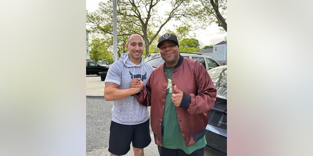 Kenan Thompson in a red jacket and green shirt holds hands with Community Officer Frank Pirri
