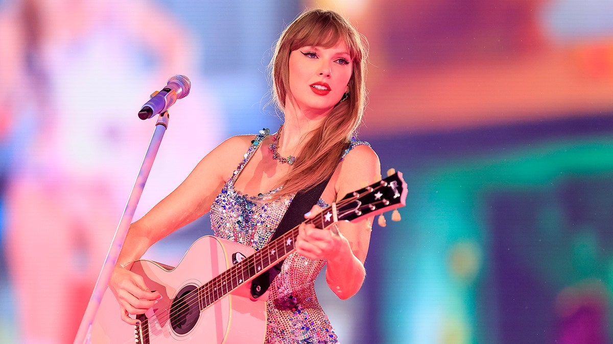 Taylor Swift in a multi colored sparkly body suit strums the guitar on stage at the Eras Tour