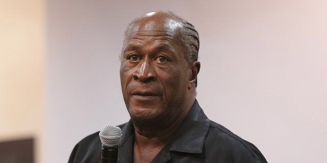 John Amos holds a microphone while speaking to a crowd
