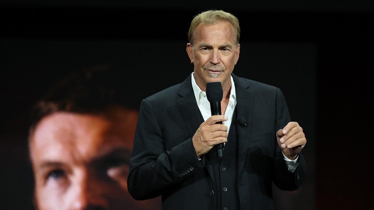 Costner talks on stage wearing a black suit holding a microphone