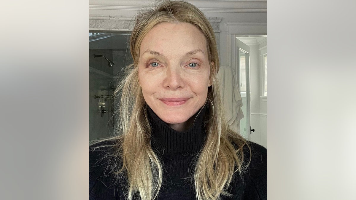 Michelle Pfeiffer in a black turtleneck stands in her bathroom and shows off her swollen eye