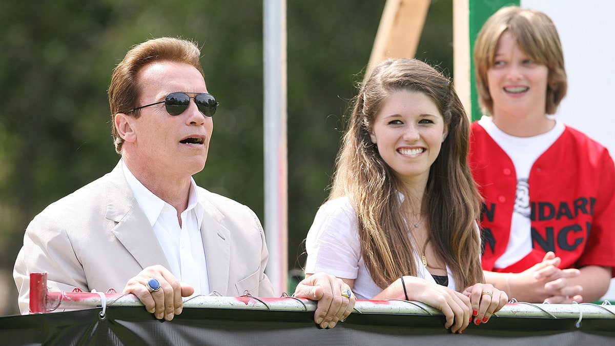 Arnold Schwarzenegger in a light tan suit and white shirt looks over the fence at a baseball game with his children Katherine and Patrick when they were young